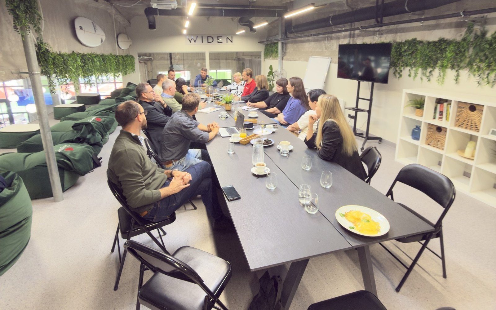 The full brunch table with nearly 20 guests in a bright room with plants and a screen